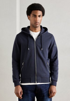 Sudadera con cremallera en color azul marino, con capucha ajustable, dos bolsillos laterales, y puños y dobladillo acanalados. Presenta una textura suave y un sutil logotipo en la manga.