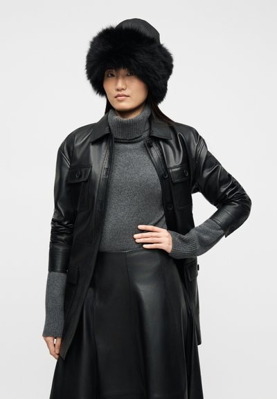 Γυναίκα που φοράει μαύρο γούνινο καπέλο, μαύρο δερμάτινο τζάκετ, γκρι ζιβάγκο πουλόβερ και μαύρη δερμάτινη φούστα, στέκεται με το ένα χέρι στη μέση.
