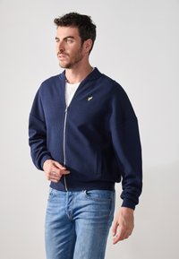 Chaqueta bomber en color azul marino con un patrón de punto texturizado, puños y dobladillo acanalados, cremallera completa y un pequeño logo dorado en el lado izquierdo del pecho. Combinada con jeans claros.