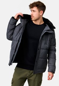 Chaqueta tipo puff en gris oscuro con un cuello de piel sintética, cierres de cremallera y broche, puños acanalados y textura acolchada. Lleva puesta una sudadera negra.