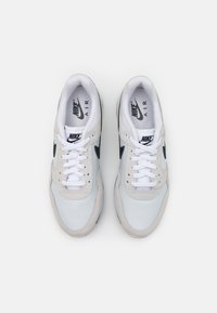 Sapatilhas Nike brancas e cinza claro com materiais em malha e camurça, detalhes pretos e atacadores brancos planos. Apresenta uma unidade de ar visível.