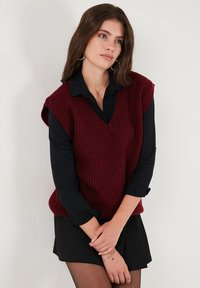 Burgundowa dzianinowa kamizelka z dekoltem w serek i wycięciami na ramionach, noszona na czarnej bluzce z długim rękawem, zestawiona z czarną mini spódnicą.