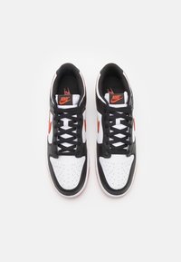 Svartvitt sportskor med orange detaljer, utrustad med perforerat tåskydd, platta svarta skosnören och gummisulor.