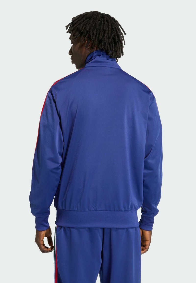 Blazer in het blauw gemaakt van gladde stof, met een opstaande kraag, lange mouwen en rode zijstrepen langs de armen.