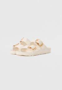 Sandali aperti in materiale color crema, con due ampie cinghie dotate di fibbie in tonalità oro e suola texturizzata per una buona aderenza.