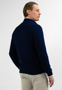 Man met licht haar in een marineblauwe kabeltrui en beige broek, zijwaarts gericht tegen een eenvoudige lichte achtergrond.