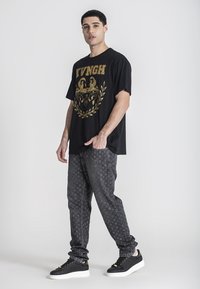 Camiseta negra con diseño gráfico en oro, combinada con pantalones grises estampados y zapatillas negras con suelas blancas y acentos dorados.