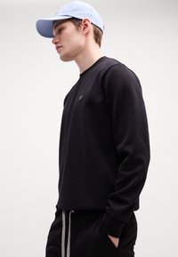 Sort sweatshirt med lange ærmer og rund halsudskæring, prydet med et lille logo på brystet. Kombinationen inkluderer en lyseblå kasket.