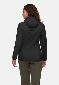 Kvinna med lockigt hår som bär en svart huva-jacka och olivgrön byxor, vänd bort från kameran mot en enkel ljus bakgrund.