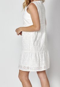 Robe midi blanche sans manches en broderie anglaise avec découpes florales, coupe ample et ourlet à volants portée par une personne debout de côté.