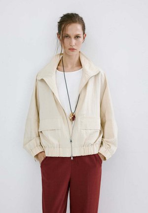 Mujer con chaqueta beige con cremallera sobre camisa blanca, pantalones rojo oscuro y un collar largo negro con colgantes dorados y rojos.