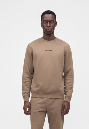 Homme portant un sweat-shirt Björn Borg marron clair et un pantalon assorti, debout devant un fond blanc uni.