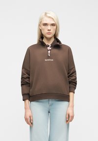 Ξανθιά γυναίκα με καφέ φούτερ με φερμουάρ στο ένα τέταρτο και λογότυπο "NAPAPIJRI" και ανοιχτόχρωμο μπλε τζην, στέκεται μπροστά σε λευκό, απλό φόντο.