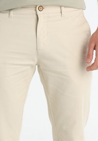 Pantalons slim fit beiges en tissu mélange coton, avec une fermeture à bouton à l'avant et une texture subtile. Pas de motifs ni d'accents visibles.