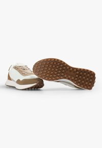 Beżowe i białe sneakersy z materiałów zamszowych i siatkowych, o zaokrąglonych noskach, z teksturowaną brązową podeszwą i wytłoczonym logo na zewnętrznej stronie podeszwy.