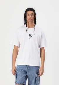 T-shirt bianco in cotone con un design di rosa nera al centro. Caratterizzato da una vestibilità comoda e maniche corte, abbinato a pantaloni cargo in denim.