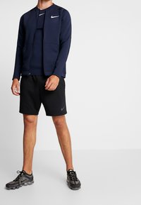Marine blå zip-up jakke med lommer, båret over en mørk top og sorte shorts, og parret med sorte sportssko. Minimalistisk design og logoer.