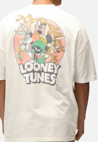 Bílý bavlněný tričko s barevným grafickým motivem Looney Tunes na zadní straně, zobrazující několik kreslených postaviček v kruhovém designu.