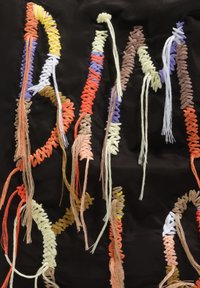 Dzieło tekstylne przedstawiające kolorowe wzory z nici na czarnym tle, z poskręcanymi i splecionymi przędzami w kolorach pomarańczowym, żółtym, fioletowym i kremowym.