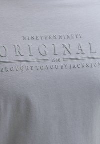 Grauer Stoff mit erhabener Schrift, auf der "Nineteen Ninety Original 1990 Brought to you by Jack & Jones" steht, zentriert auf der Oberfläche.