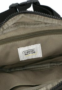 Interior de bolsa de tela gris con múltiples compartimentos con cremallera, herrajes negros y una etiqueta visible de "Camel Active". Textura suave.