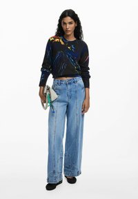 Pull noir avec des motifs abstraits colorés, accompagné d'un jean en denim bleu clair à jambes larges. Le mannequin tient une petite pochette grise.
