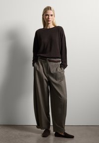Maglione a maglia marrone scuro, pantaloni grigi a pieghe a gamba larga, cintura in pelle marrone e scarpe scure. Tessuto liscio, vestibilità rilassata, design minimale.