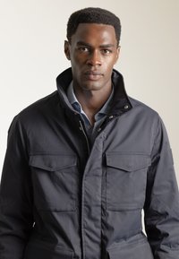 Chaqueta azul marino con un cuello alto, dos bolsillos en el pecho y cierre de cremallera en la parte delantera. Presenta una textura lisa y mate, con detalles de botones.