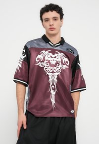 Μπλούζα μπορντό και γκρι με μαύρες λεπτομέρειες, διακοσμημένη με λευκό φυλετικό μοτίβο. Κατασκευασμένη από γυαλιστερό ύφασμα, με κοντά μανίκια και γιακά.