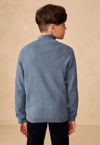 Strikket blå sweater med høj krave og lange ærmer. Har en glat struktur og ribstrikkede manchetter. Ren rygdesign uden synlige mønstre.
