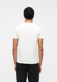 Άνδρας με κοντά σγουρά μαλλιά που φοράει απλό λευκό t-shirt με κοντά μανίκια και μαύρο παντελόνι, γυρισμένος από την πλάτη σε απλό φόντο.