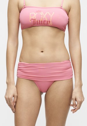 Femme portant un maillot de bain deux pièces rose avec le texte « ROXY Juicy » sur le haut et des détails froncés sur le bas, montrée des épaules aux cuisses.