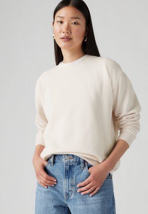Femme aux cheveux raides et foncés, portant des boucles d'oreilles anneaux dorées, un pull crème à manches longues et col rond, et un jean bleu taille haute avec les mains dans les poches.