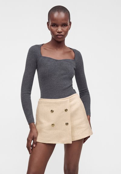 Femme portant un top gris côtelé à manches longues ajusté et une mini-jupe beige texturée taille haute avec des boutons dorés sur un fond uni.