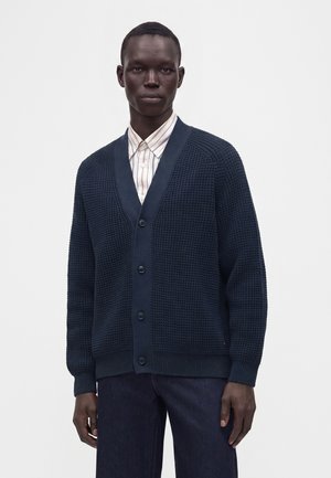 Man draagt een donkerblauwe gestructureerde cardigan met knopen over een licht gestreept overhemd en een donkere broek tegen een effen witte achtergrond.