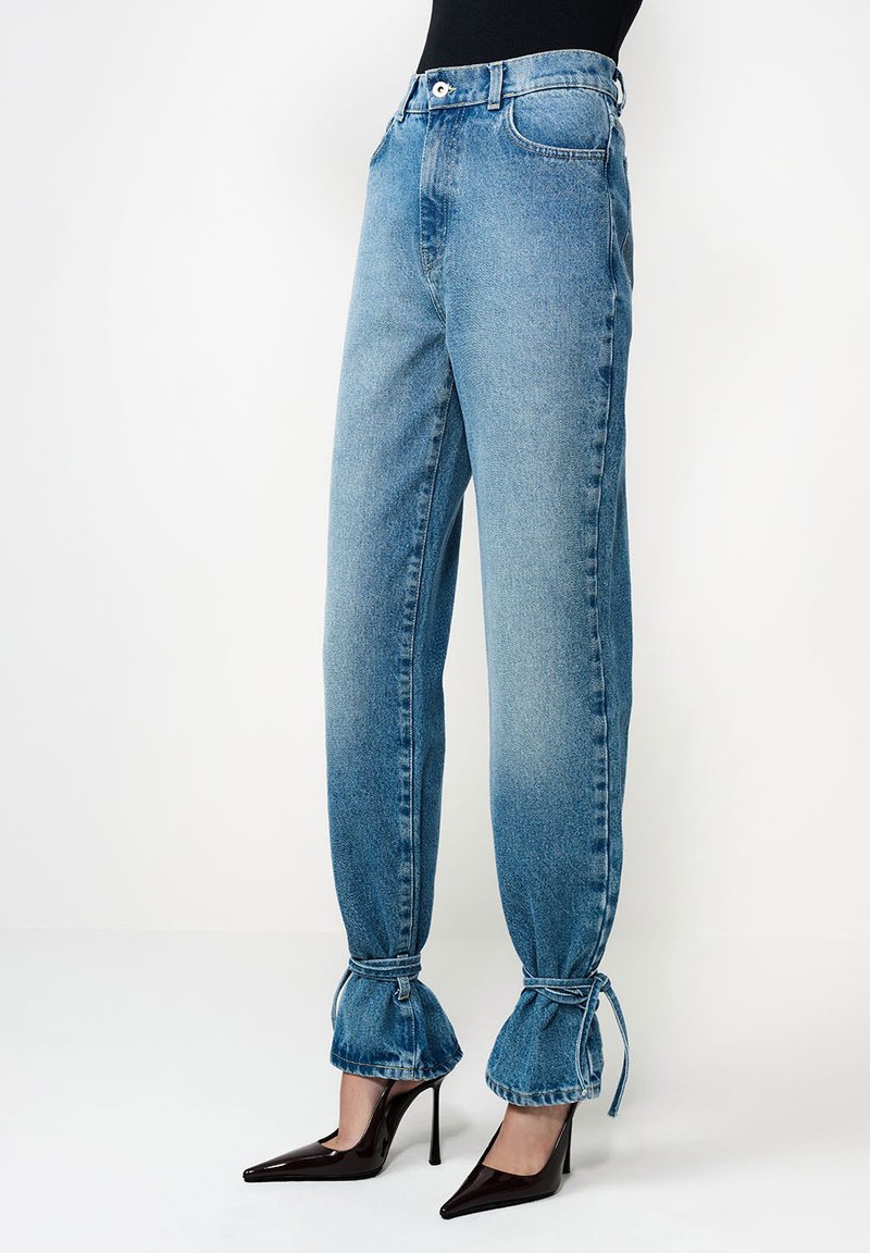 Blugi din denim albastru deschis, cu croială conică, șnururi strânse la gleznă și închidere clasică cu nasturi și fermoar. Asortați cu pantofi negri cu toc ascuțit.