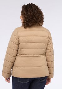 Beige pufferjacka med quiltat mönster, rundad krage och textilmaterial. Har elastiska muddarna och en figursydd siluett.