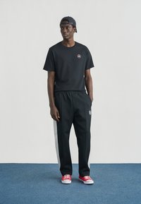 Schwarzes Baumwoll-T-Shirt mit Logopatch, kombiniert mit schwarzen Jogginghosen mit weißen Seitenstreifen. Rote Turnschuhe vervollständigen das Outfit.