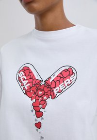 Close-up van een wit shirt waarop een open capsule te zien is die rode harten en zilveren hartvormige embellishments morst, met gedeeltelijke tekst "SCAL" en "PERS".