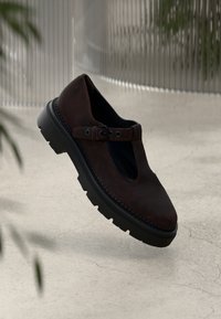 Chaussure en cuir marron foncé avec un design découpé, une sangle à boucle ajustable et une semelle en caoutchouc noir épais avec une texture de semelle.