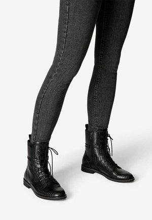 Jambes vêtues d’un jean gris foncé ajusté et de bottines noires texturées à lacets avec fermetures éclair latérales sur un fond clair.