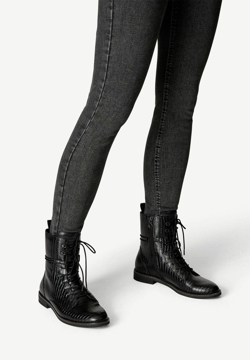 Jambes vêtues d’un jean gris foncé ajusté et de bottines noires texturées à lacets avec fermetures éclair latérales sur un fond clair.