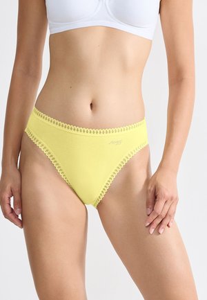 Vrouw in gele bikini-onderbroek met kant en een witte beha tegen een effen lichte achtergrond.