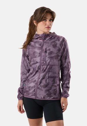 Vrouw draagt een paarse zip-up jas met camouflerend patroon en capuchon, en zwarte fietsshorts, staand tegen een effen achtergrond.