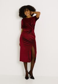 Robe midi rouge à manches courtes, détail noué froncé à la taille, fente sur le côté gauche, confectionnée en tissu doux et texturé avec un léger éclat.
