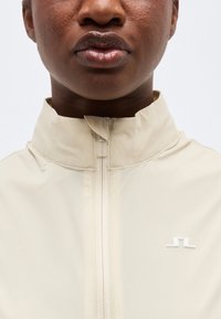 Κοντινό πλάνο ενός ατόμου που φοράει μπεζ μπουφάν με φερμουάρ και ψηλό γιακά, με ένα μικρό λευκό λογότυπο στο στήθος.