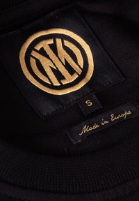 Zwarte kledinglabel met gouden cirkelvormige breiwarenlogo, maat "S" etiket en de inscriptie "Made in Europe" op zwart stof.