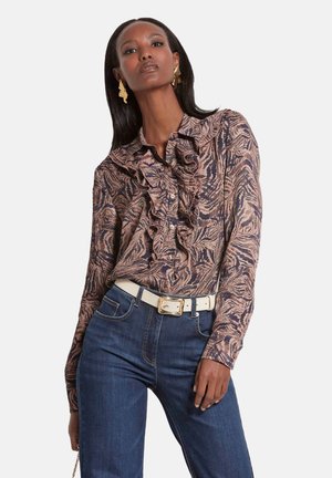 Blouse met een gerimpelde halslijn, voorzien van een abstract bruin en marineblauw patroon. Gecombineerd met een donkere spijkerbroek en een witte riem.