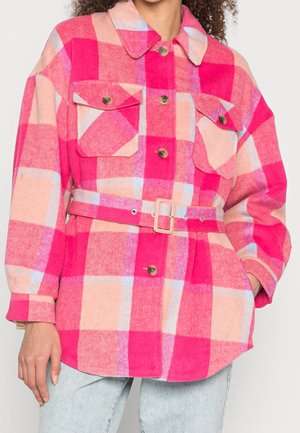 Vrouw draagt een roze en perzikkleurige geruite jas met riem en met knoopzakken op de borst, gecombineerd met lichtblauwe spijkerbroeken.