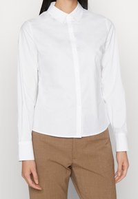 Chemise blanche boutonnée avec col pointu, manches longues et coupe ajustée, confectionnée en tissu de coton lisse. Assortie à un pantalon marron clair.
